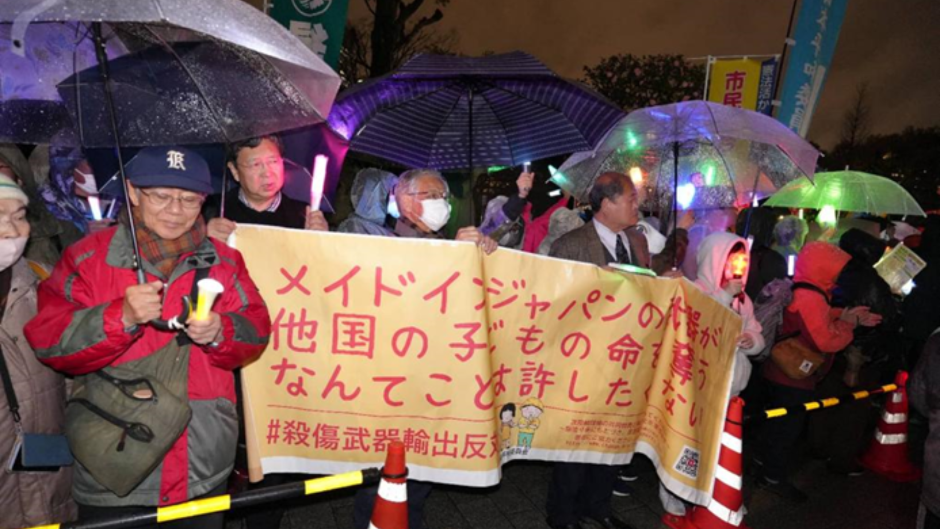 Protesters hold a banner in the rain opposing "Made in Japan" weapons exports and militarism.