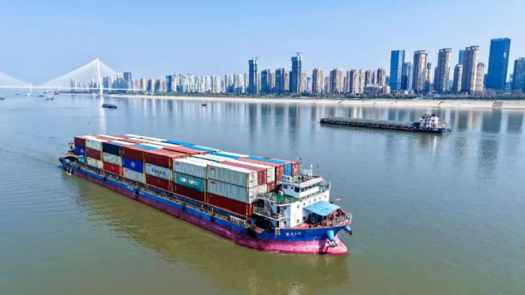 A cargo ship carrying colorful containers navigates a river toward a modern city skyline.