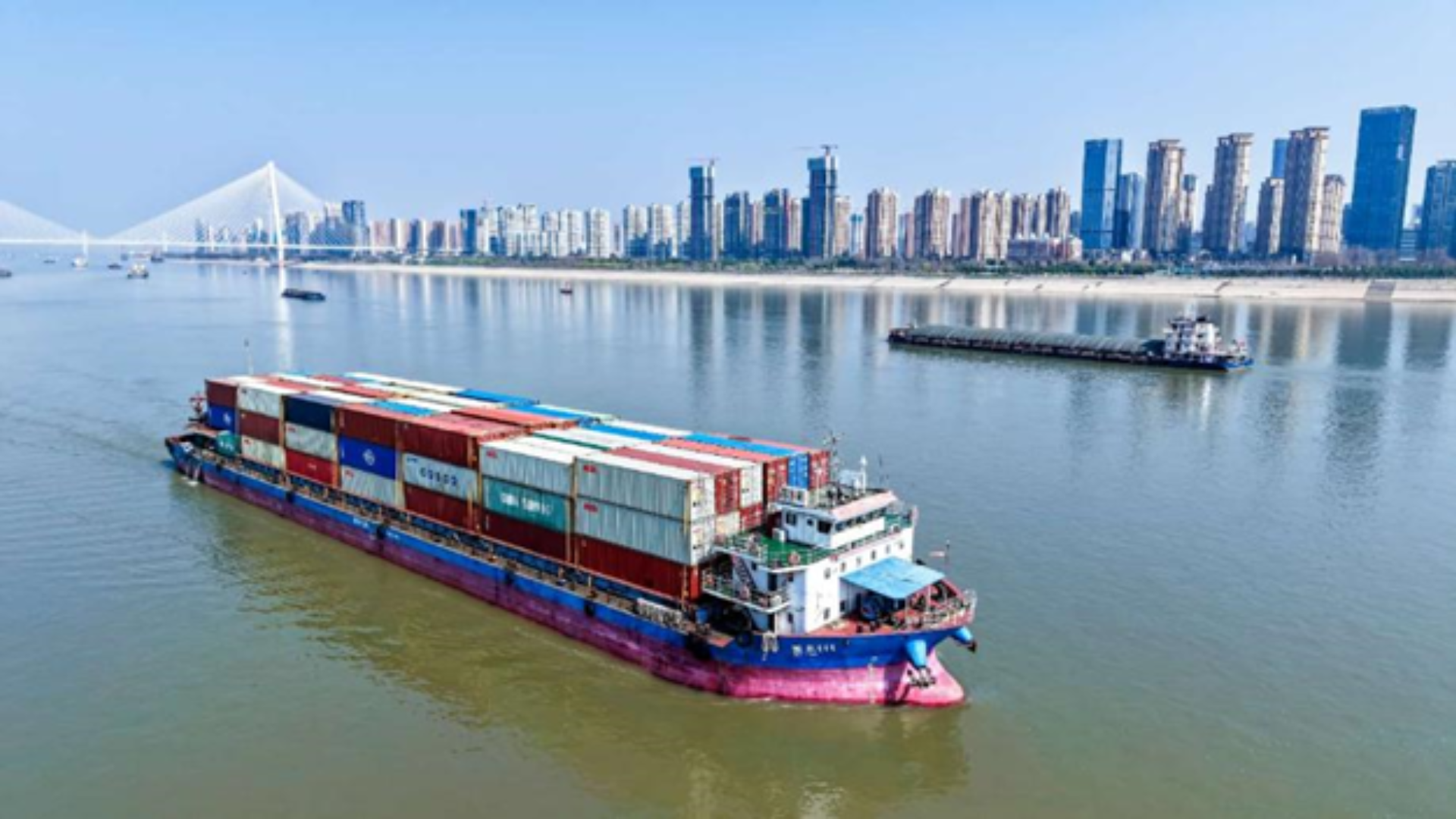 A cargo ship carrying colorful containers navigates a river toward a modern city skyline.