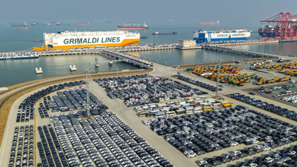 Thousands of new energy vehicles await shipment at a busy Chinese port alongside Grimaldi and COSCO vessels.