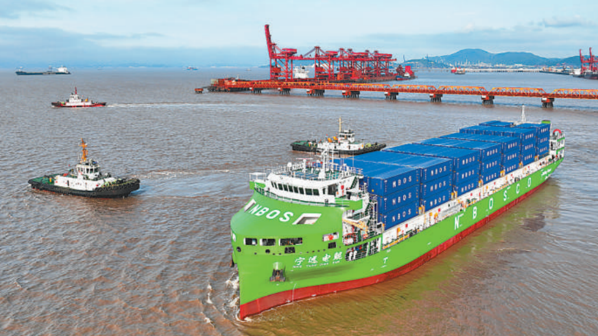 A green electric container ship sails through the sea, guided by tugboats near a port.