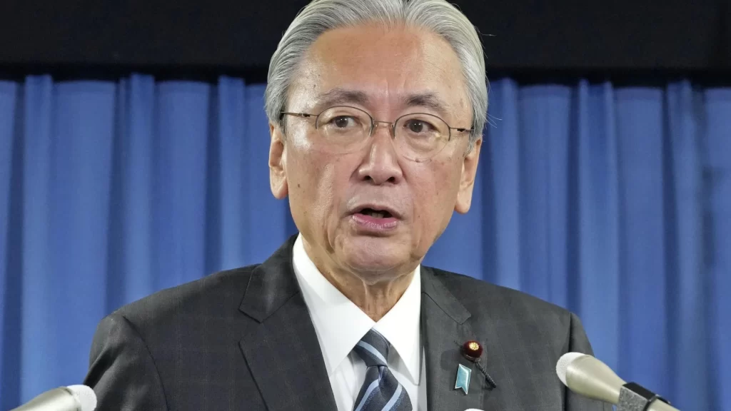 Japanese politician Keiji Furuya speaking at a microphone against a light teal background.