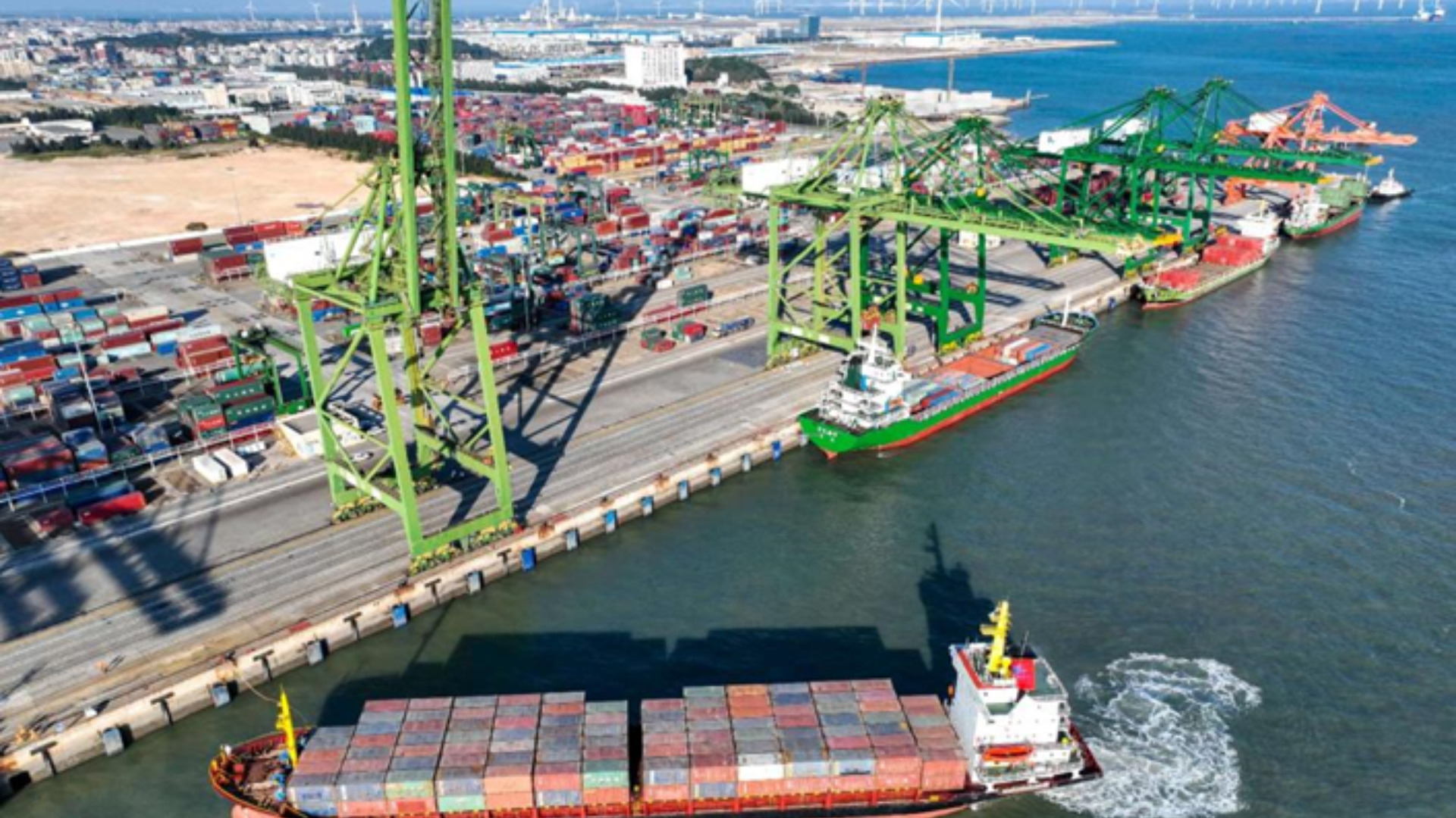 An aerial view of a busy container port featuring green cranes and a cargo ship.