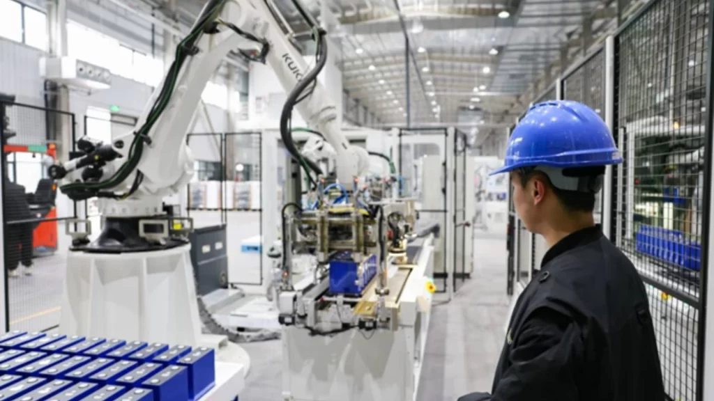 A worker in a blue hard hat monitors an automated robotic arm assembling energy storage batteries.