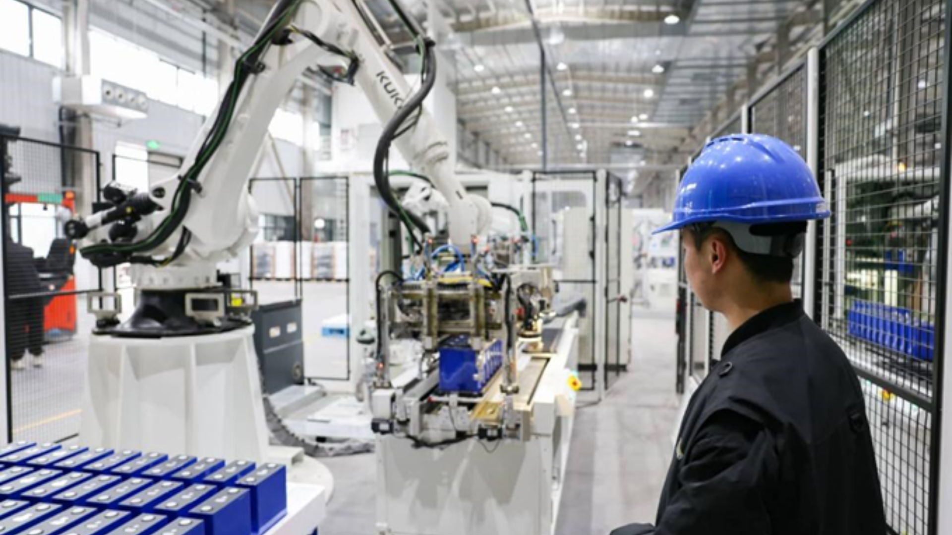 A worker in a blue hard hat monitors an automated robotic arm assembling energy storage batteries.