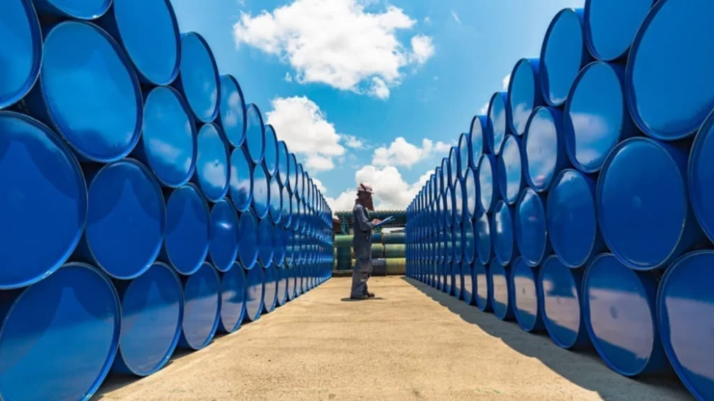 Oil worker stands between massive walls of stacked blue industrial barrels.