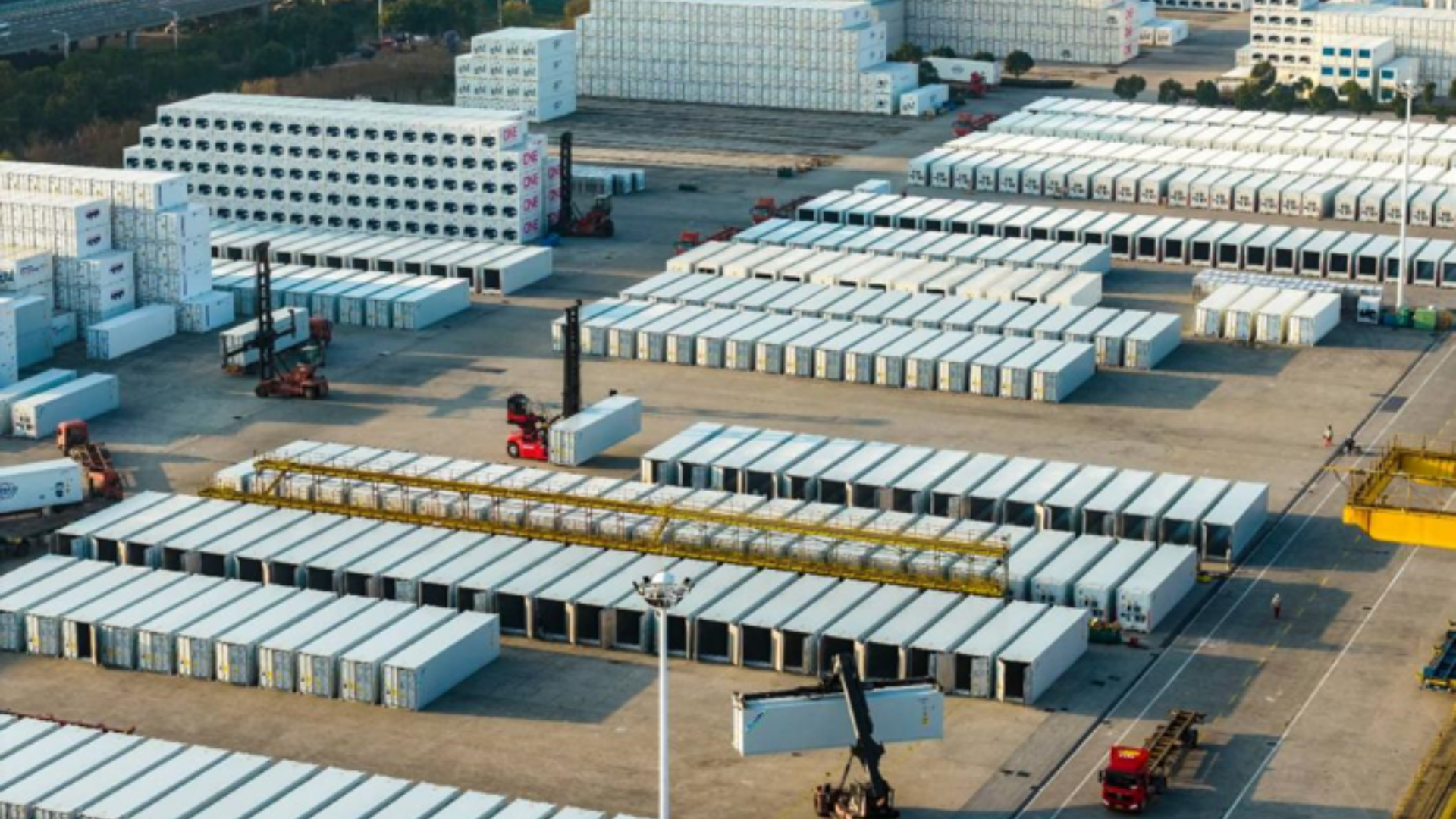 Aerial view of organized stacks of white shipping containers at a bustling industrial port facility.