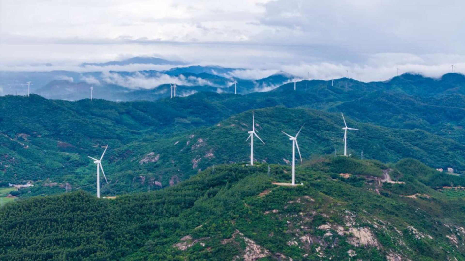 Wind turbines spin atop lush green hills under a cloudy sky in Yangjiang, China.