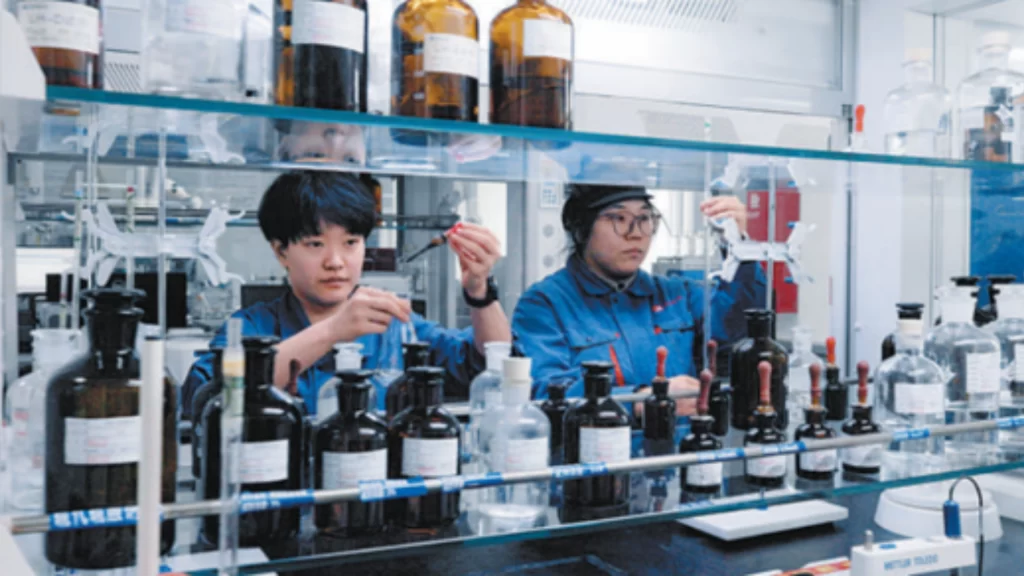 Two lab technicians inspect chemical solutions amidst glass bottles in a carbon fiber research laboratory.