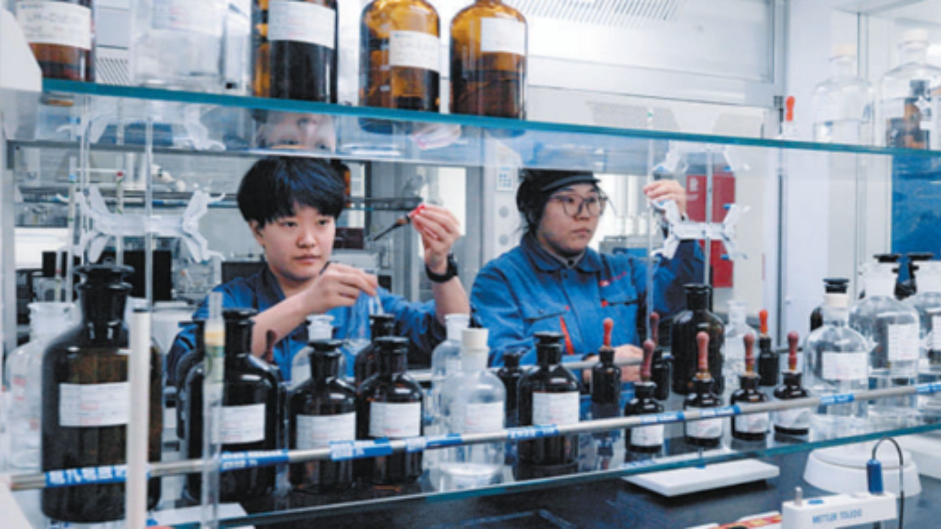 Two lab technicians inspect chemical solutions amidst glass bottles in a carbon fiber research laboratory.