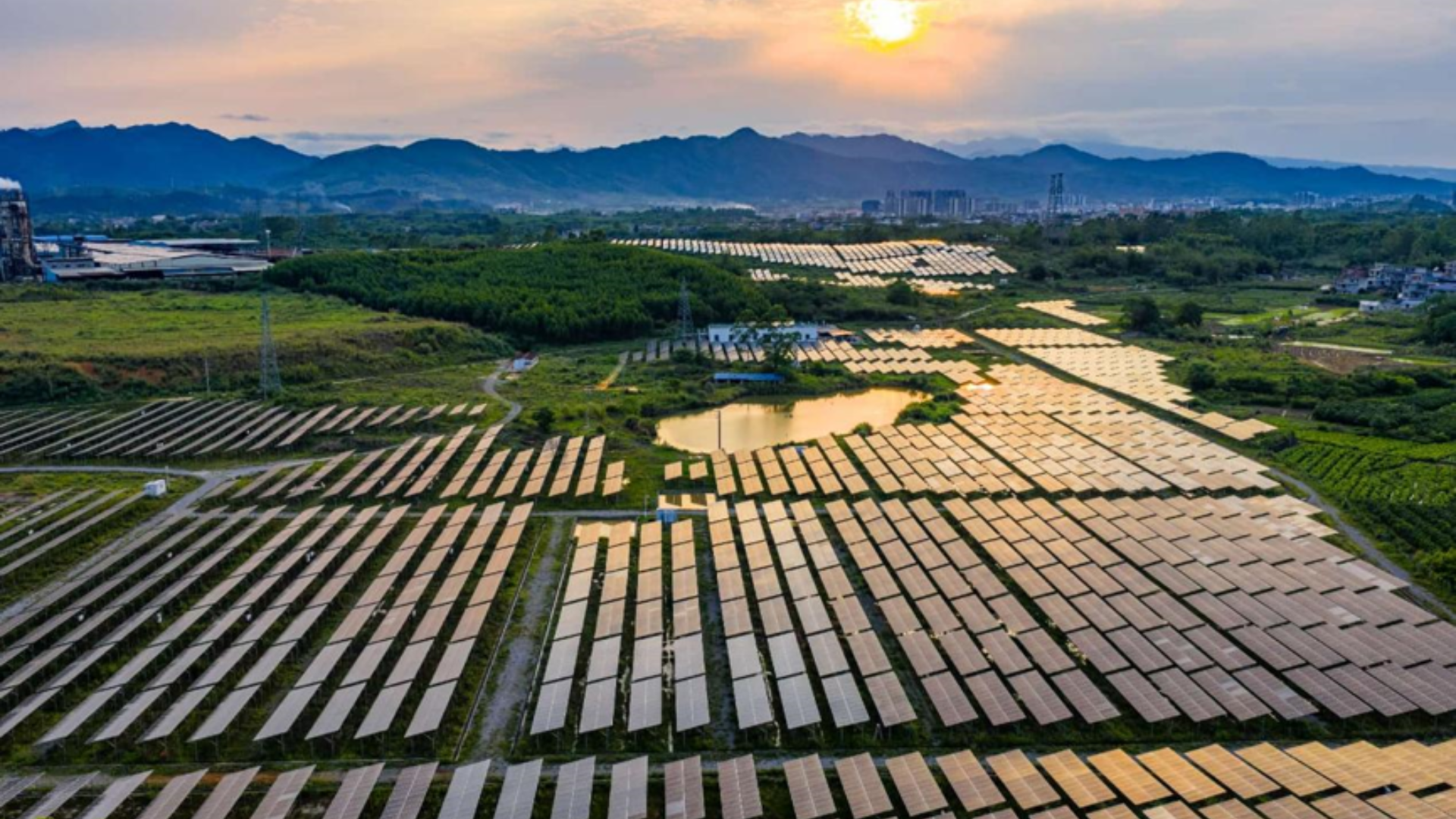 An aerial view of a massive solar panel field reflecting the setting sun over mountains.