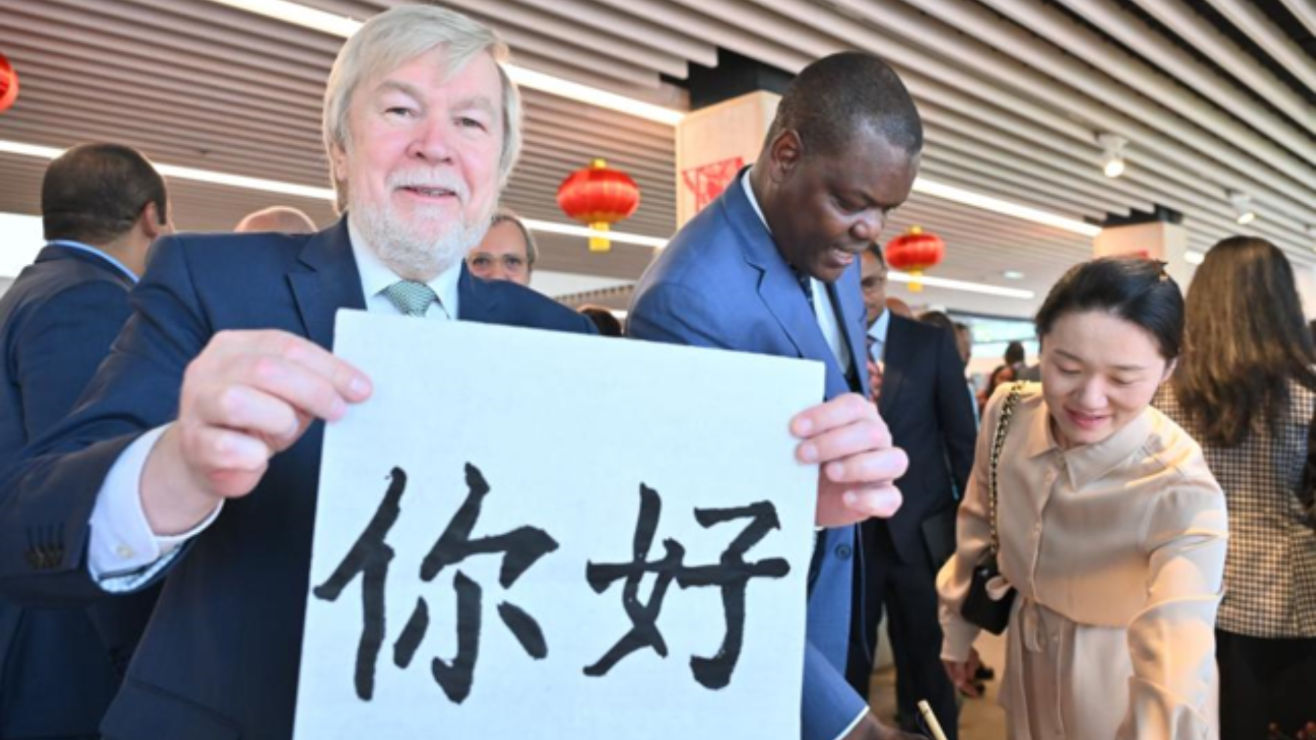 An international delegate proudly smiles while presenting a calligraphy sheet featuring the Chinese characters for "Hello."