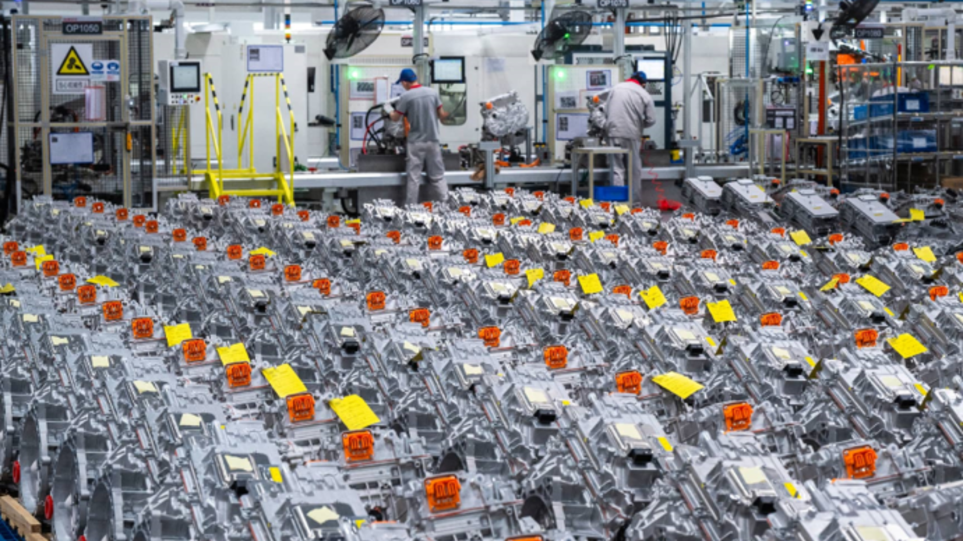 Rows of manufactured automotive components fill a factory floor with workers at assembly stations.