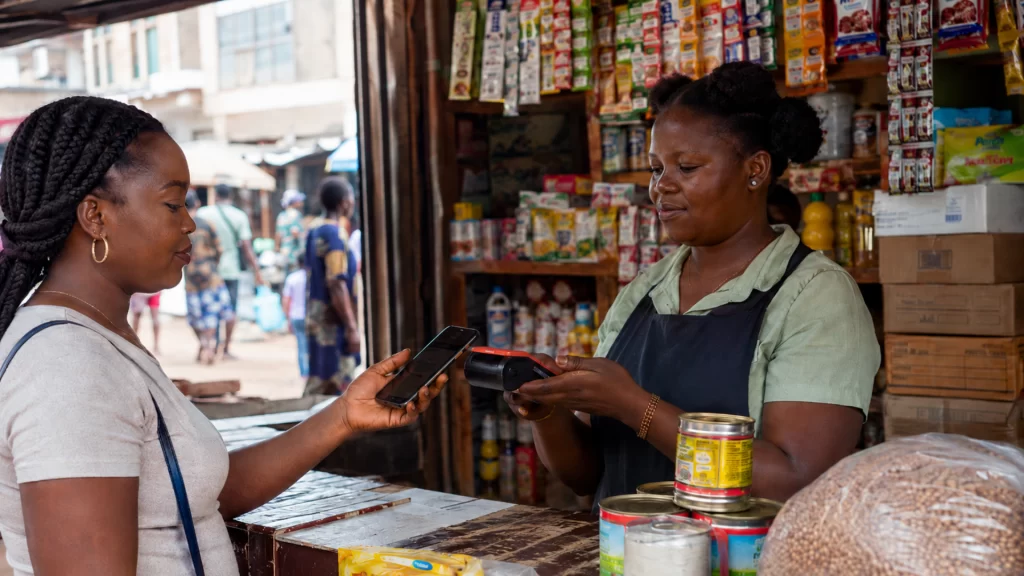 An African shopkeeper assists a customer making a cashless smartphone payment at a market.