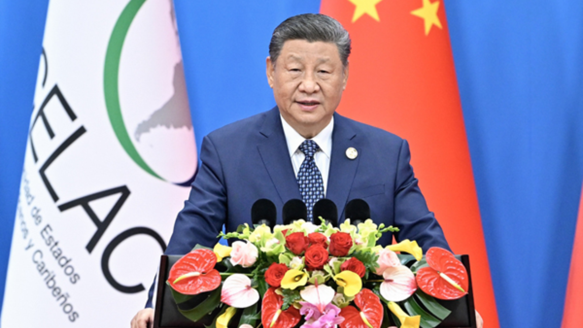 President Xi Jinping delivers a speech behind a floral arrangement and national flags.