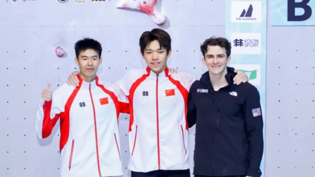 Three speed climbers stand together on a podium, smiling in front of a climbing wall.
