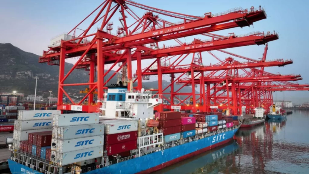 A large cargo ship loaded with containers docked beneath towering red port cranes.