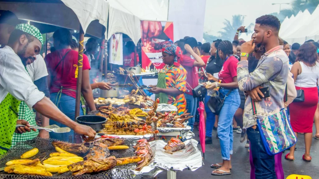 Vendors grilling plantains and fish at an outdoor street food festival with festivalgoers browsing.