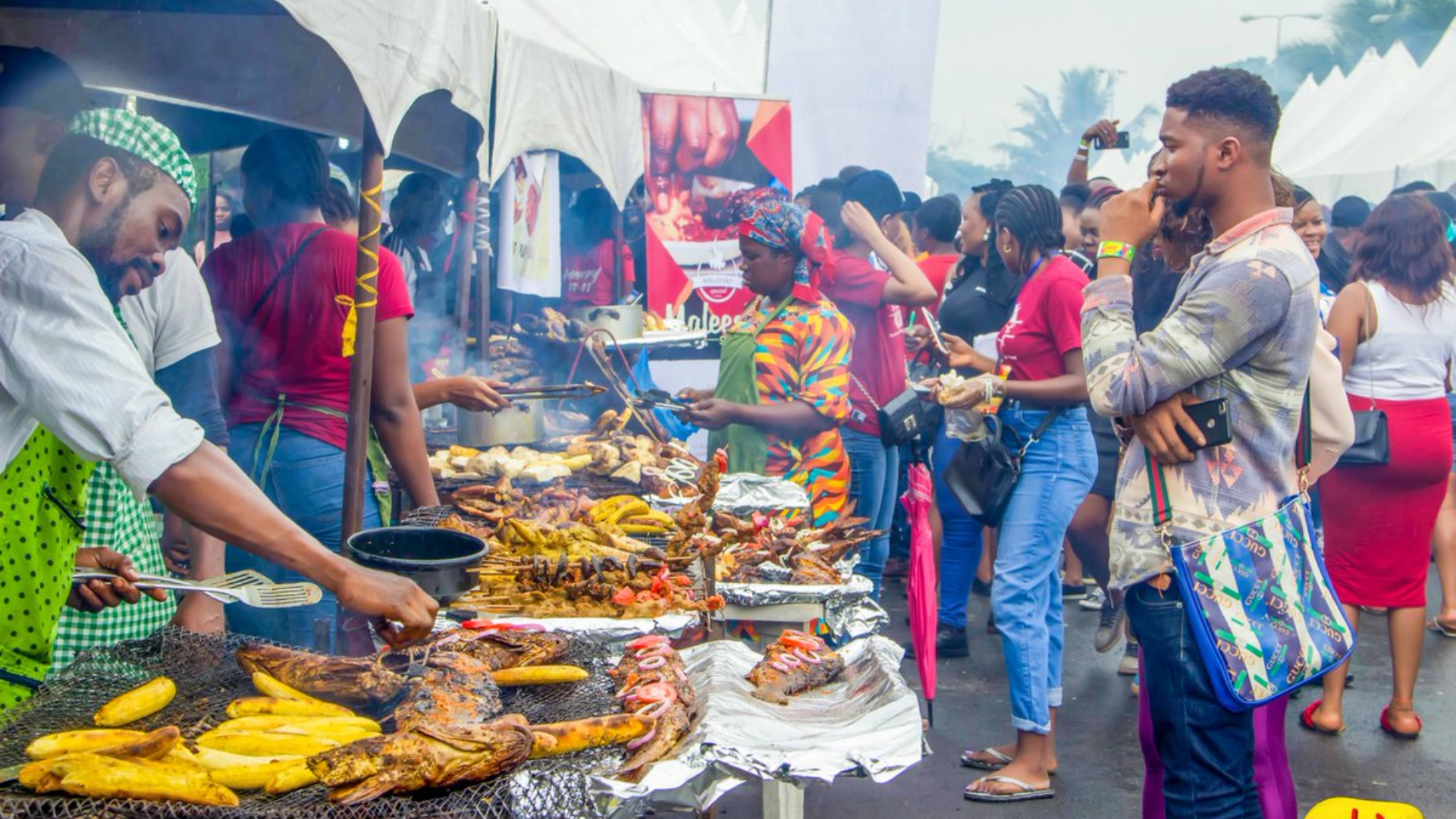 Vendors grilling plantains and fish at an outdoor street food festival with festivalgoers browsing.
