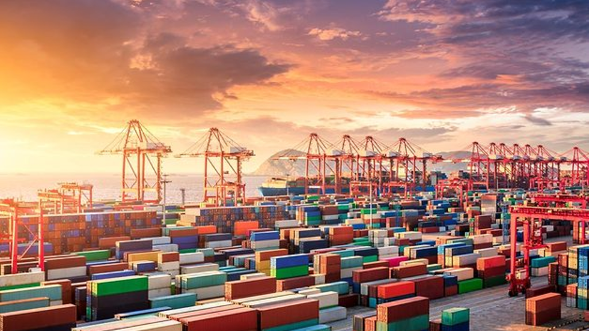A commercial shipping port with stacked containers and gantry cranes under a sunset sky.