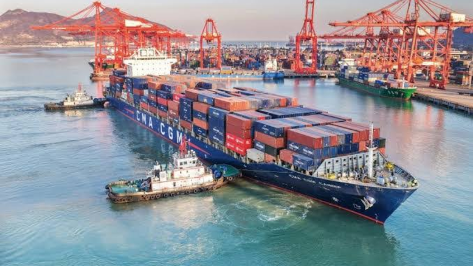 A CMA CGM container ship maneuvers through a busy port guided by tugboats.